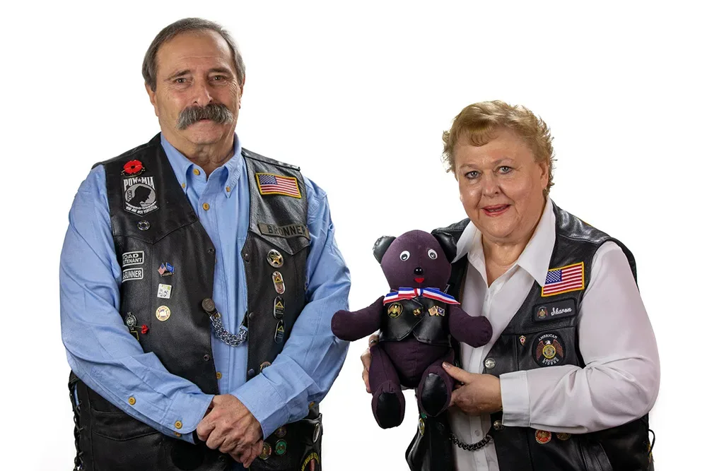 Two Riders in leather vests holding a handmade bear that wears its own miniature vest